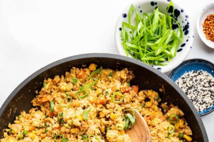 Cauliflower fried rice with eggs in a skillet with a wooden serving spoon. The dish is garnished with fresh green onion.