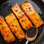 Four honey glazed salmon filets garnished with fresh chives in a skillet with a metal spoon that contains some of the honey based sauce in it.