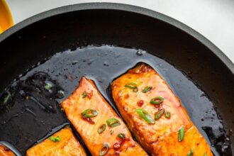 Four honey glazed salmon filets garnished with fresh chives in a skillet with a metal spoon that contains some of the honey based sauce in it.