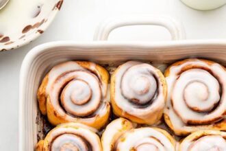 A white square baking dish filled with 9 baked cinnamon rolls that are topped with a glaze.