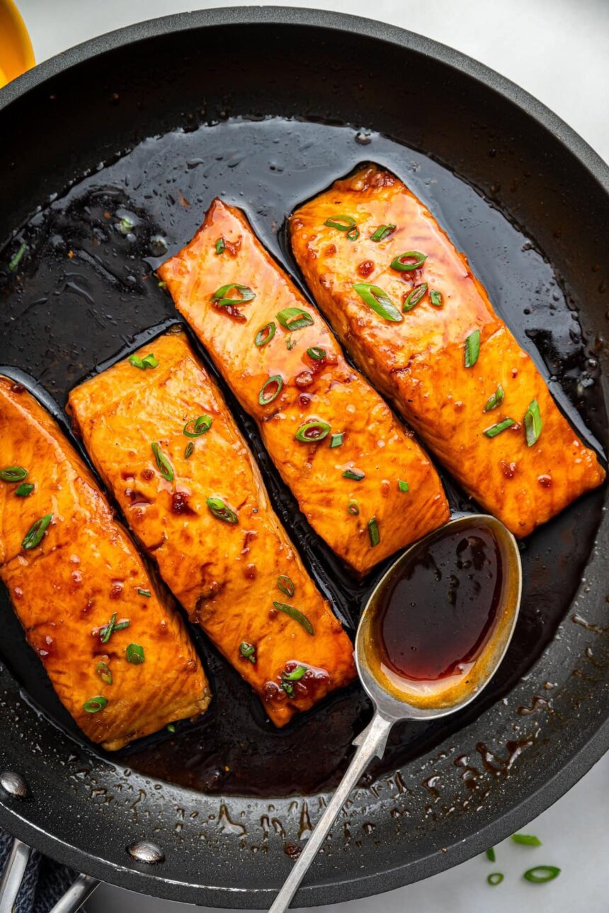 Four honey glazed salmon filets garnished with fresh chives in a skillet with a metal spoon that contains some of the honey based sauce in it.