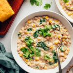 Bowls of creamy vegan white bean chili next to cayenne, cilantro, and cornbread