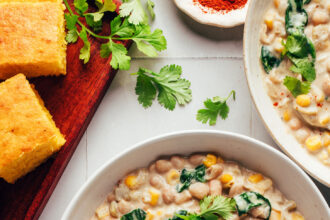 Bowls of creamy vegan white bean chili next to cayenne, cilantro, and cornbread