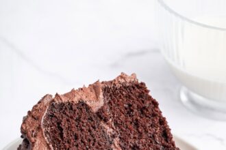 A slice of chocolate cake with chocolate buttercream frosting on a white plate with a metal fork to the side.