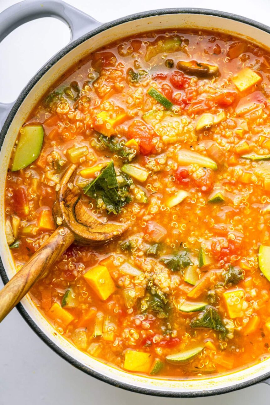 A large pot of vegetable packed quinoa soup with a wooden serving spoon in the soup.