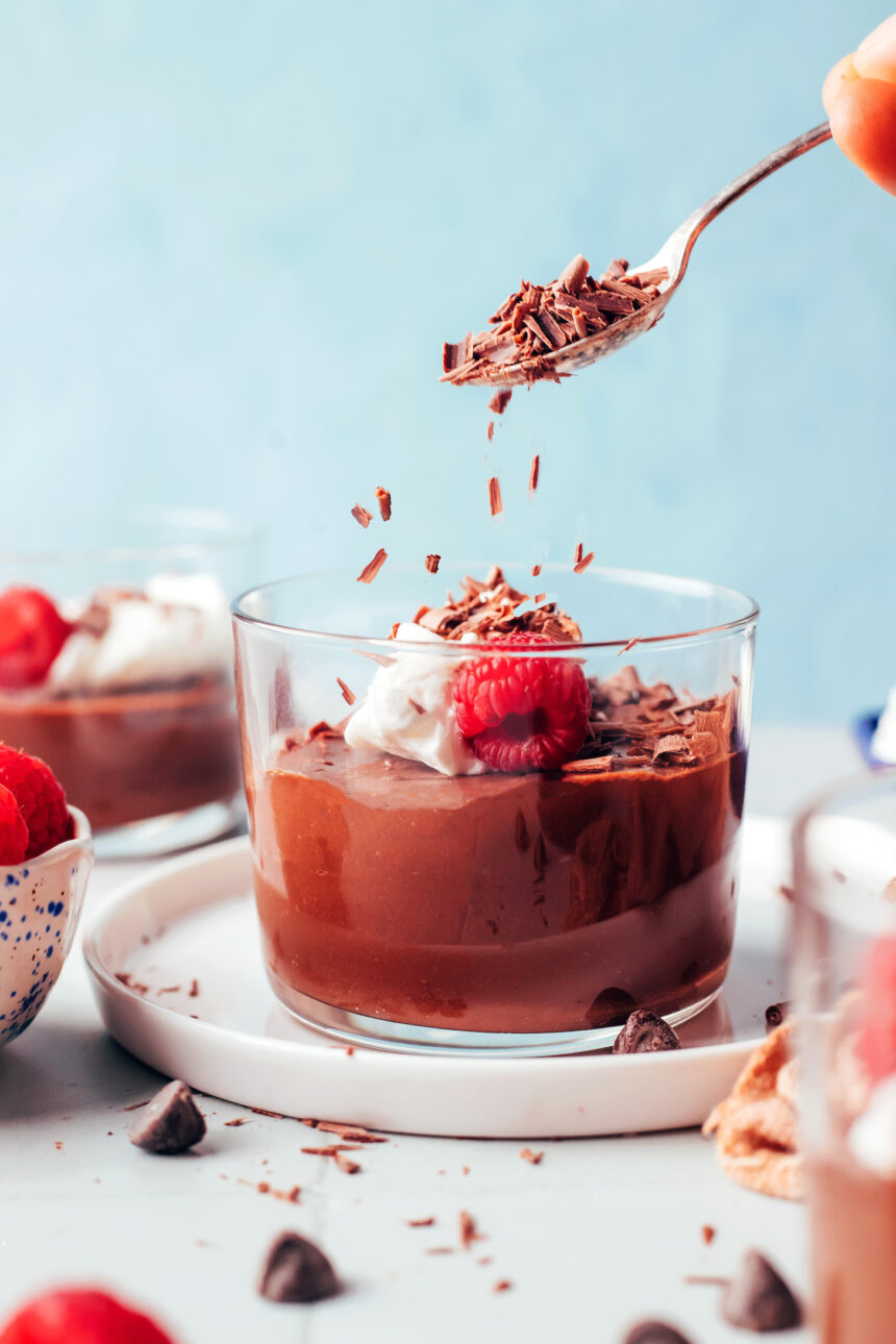 Sprinkling shaved dark chocolate into a glass of pot de creme topped with coconut whipped cream and a fresh raspberry