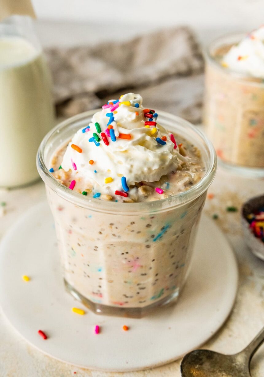 A glass jar filled with birthday cake overnight oats, topped with whipped cream and colorful sprinkles, with a bottle of milk and another jar of oats in the background.