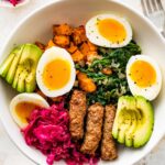 A breakfast bowl with 2 soft boiled eggs, 3 turkey sausage, sliced avocado, sauerkraut, roasted sweet potatoes and sautéed kale all in a white bowl.