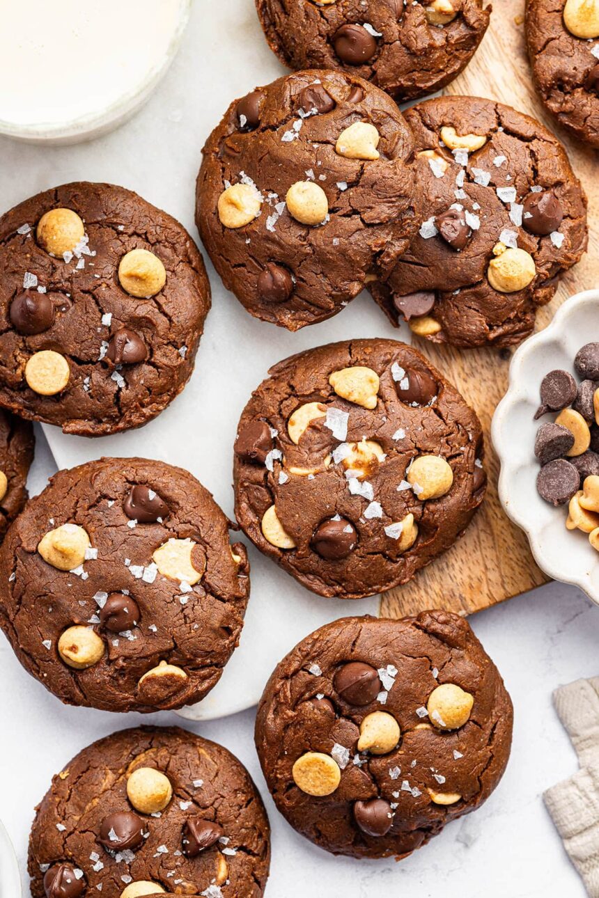 9 chocolate peanut butter cookies spread out on a counter. All of the cookies are topped with flaky sea salt.