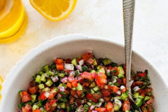 A Shirazi Salad in a white bowl served with a silver spoon. There is a lemon half and a bottle of olive olive next to the bowl.