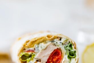 Two halves of a Greek chicken wrap stacked on top of each other on a wooden cutting board. You can see the tomatoes, chicken, lettuce and sauce inside the wrap.