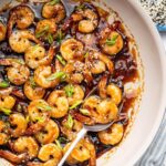 Shrimp coated in a honey garlic sauce in a skillet. There is a silver spoon in the skillet and the shrimp is topped with sesame seeds and green onion slices.