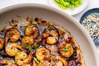 Shrimp coated in a honey garlic sauce in a skillet. There is a silver spoon in the skillet and the shrimp is topped with sesame seeds and green onion slices.