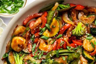 Shrimp stir-fry with shrimp, broccoli, red bell peppers, snap peas, zucchini, and onions coated in sauce. The dish is garnished with sesame seeds and chopped green onions, with a wooden spoon in the pan.