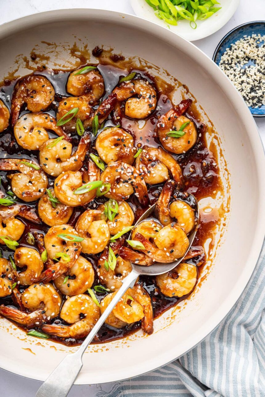 Shrimp coated in a honey garlic sauce in a skillet. There is a silver spoon in the skillet and the shrimp is topped with sesame seeds and green onion slices.