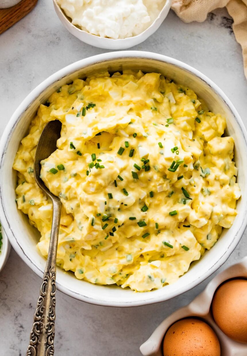 Cottage cheese egg salad served in a white ceramic bowl served with a silver spoon. Fresh chives are topping the egg salad.