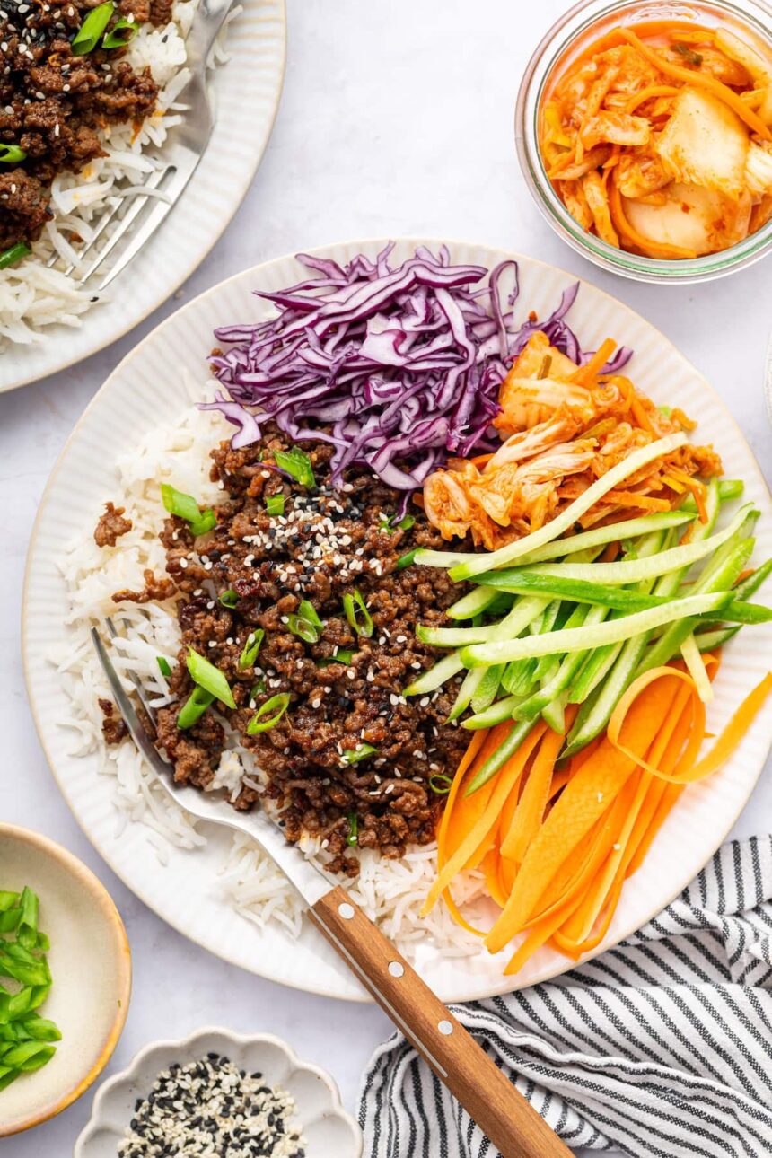 Korean beef served over white rice with fresh carrots, purple cabbage, cucumber and kimchi. The beef is topped with sesame seeds and green onions.