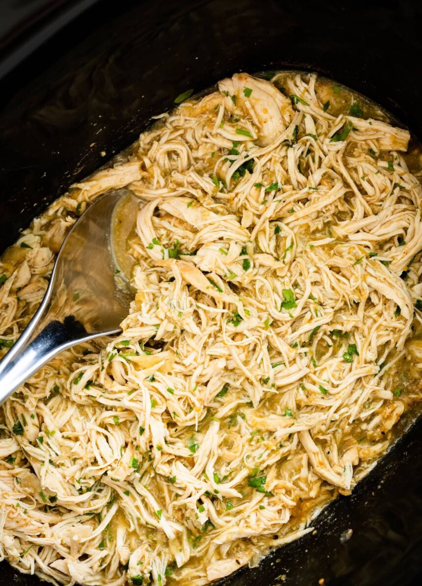 Shredded salsa verde chicken in a slow cooker with a silver serving spoon. Fresh cilantro is on top of the chicken.