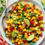 Fork and spoon resting in a bowl of Mango Cucumber Salad with lime wedges on top