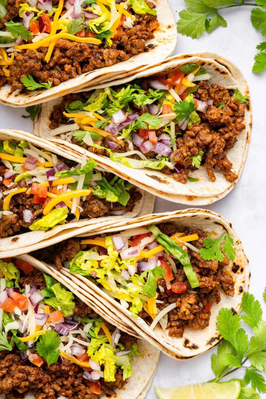 Five ground beef tacos in flour tortillas topped with lettuce, onion, tomato, cilantro and cheese.