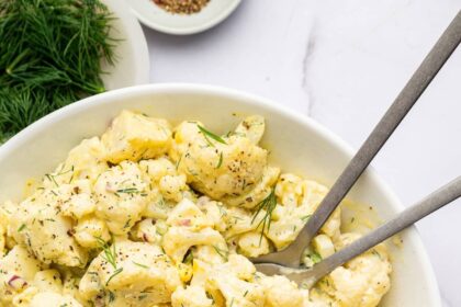 Cauliflower potato salad in a large serving bowl with silver serving utensils. The salad is topped with fresh dill.