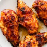 Four grilled BBQ chicken breasts on a white serving plate.