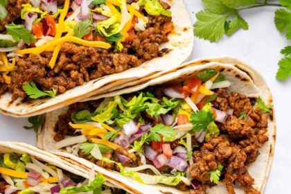 Five ground beef tacos in flour tortillas topped with lettuce, onion, tomato, cilantro and cheese.