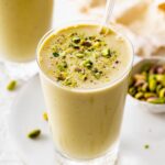 One glass filled with a pistachio smoothie topped with crushed pistachios. There is a clear straw in the glass and another smoothie in the background.