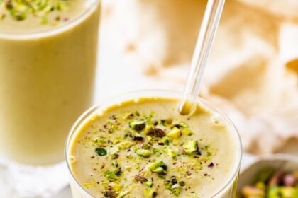 One glass filled with a pistachio smoothie topped with crushed pistachios. There is a clear straw in the glass and another smoothie in the background.