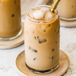 A glass of iced coffee with milk fully mixed in, served on a small wooden plate with a metal straw.
