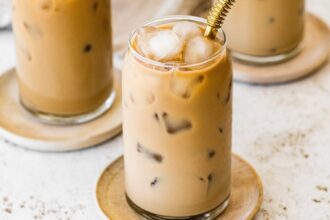 A glass of iced coffee with milk fully mixed in, served on a small wooden plate with a metal straw.