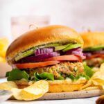 A chicken burger served on a brioche bun topped with lettuce, tomato, red onion and avocado. The burger is served with a side of potato chips.