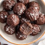 Bowl of chocolate protein balls stacked with flaky sea salt on top and one bitten into.