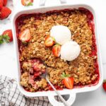 Baked strawberry crisp topped with two scoops of vanilla ice cream and fresh strawberry halves. There is a spoon scooping out some of the crisp.