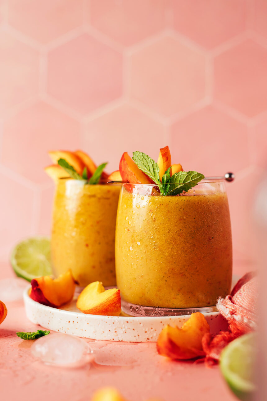 Two glasses of our frozen peach daiquiri on a plate with peach slices