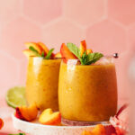 Two glasses of our frozen peach daiquiri on a plate with peach slices