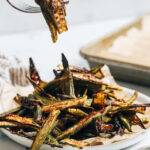 Fork holding up a few slices of crispy oven roasted okra over a plate of more baked okra
