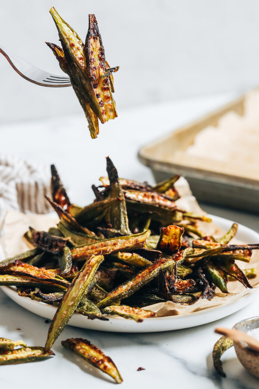 Fork holding up a few slices of crispy oven roasted okra over a plate of more baked okra