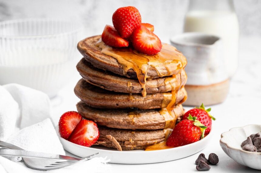 Tall stack of chocolate protein pancakes topped with fresh strawberries and nut butter drizzle.