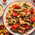 Large serving bowl of garlicky pasta with burst cherry tomatoes, kale, olives, and vegan parmesan cheese