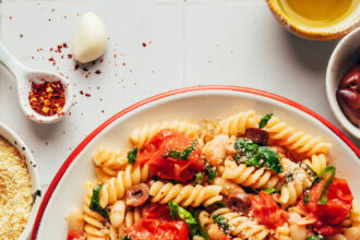 Large serving bowl of garlicky pasta with burst cherry tomatoes, kale, olives, and vegan parmesan cheese