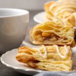 Two halves of an apple turnover stacked on a plate with a coffee cup and more turnovers in the background.
