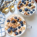 Two glass bowls of overnight oats topped with fresh blueberries and walnuts, set on a white table with gold spoons and a blue-and-white napkin.