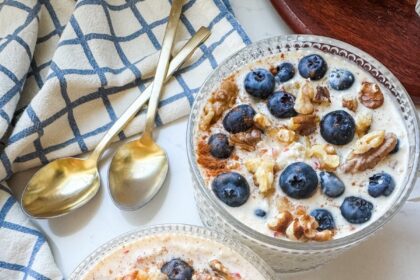 Two glass bowls of overnight oats topped with fresh blueberries and walnuts, set on a white table with gold spoons and a blue-and-white napkin.