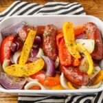 A close up shot of a baking dish filled with sausage and peppers with onions, over a black and white striped tea towel on a wooden tabletop.