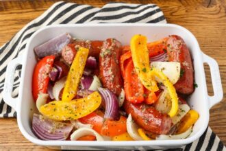 A close up shot of a baking dish filled with sausage and peppers with onions, over a black and white striped tea towel on a wooden tabletop.