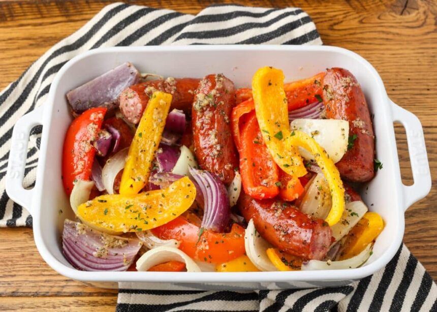 A close up shot of a baking dish filled with sausage and peppers with onions, over a black and white striped tea towel on a wooden tabletop.