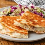 You can see chunks of tender turkey in this close up horizontal shot of a turkey quesadilla on a white plate over a wooden tabletop with a grey and white checkered tea towel in the background.
