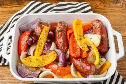 A close up shot of a baking dish filled with sausage and peppers with onions, over a black and white striped tea towel on a wooden tabletop.