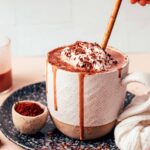 Small bowl of cocoa powder next to an overflowing mug of dairy-free tahini hot chocolate